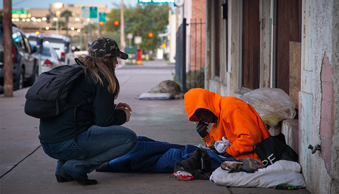 Person kneeling and talking with a homeless individual sitting on a sidewalk.